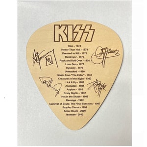 May include: A wooden guitar pick with a brown background and the word "KISS" in large, bold letters. The pick lists the band's albums and release years, including "Kiss", "Hotter Than Hell", "Dressed to Kill", "Destroyer", "Rock and Roll Over", "Love Gun", "Dynasty", "Unmasked", "Music from "The Elder"", "Creatures of the Night", "Lick It Up", "Animalize", "Asylum", "Crazy Nights", "Hot in the Shade", "Revenge", "Carnival of Souls: The Final Sessions", "Psycho Circus", "Sonic Boom", and "Monster".