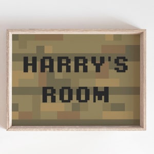 May include: A framed print with the text "HARRY'S ROOM" in pixelated black letters. The background features a camouflage pattern in shades of brown and green. The frame is a light wood colour.