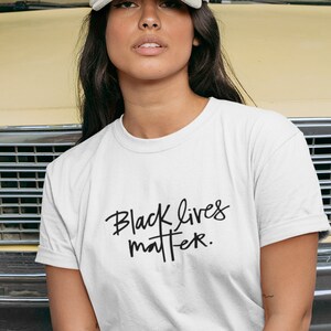 May include: A person wearing a white t-shirt with the text "Black Lives Matter" in black script. The person is wearing a grey baseball cap and is sitting in front of a yellow car.