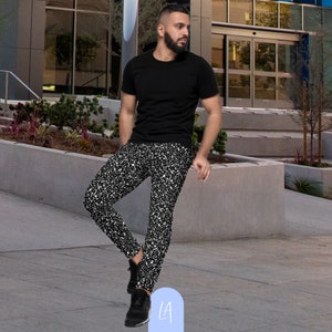 May include: A person wearing a black t-shirt and black and white patterned pants. The pants have a white and black speckled pattern. The person is standing on a sidewalk in front of a building.