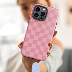 May include: A pink and light pink chequered phone case. The case is held in a person's hand, showing the design. The phone case has a black border and a camera cutout.