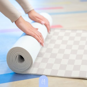 Peut inclure: Une personne enroule un tapis de yoga blanc avec un motif à carreaux sur un sol en bois clair.