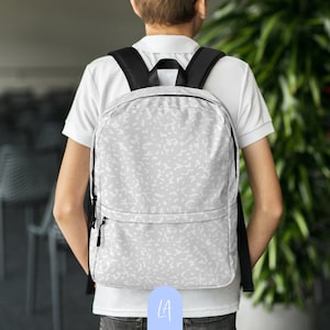 Puede incluir: Una mochila blanca con un patrón abstracto blanco y negro. La mochila tiene una cremallera negra y una correa ajustable negra.