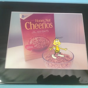 May include: A box of Honey Nut Cheerios cereal with a cartoon bee mascot standing in front of a bowl of cereal. The box is red and white with the words "Honey Nut Cheerios" and "Whole Grain Oats". The bee is yellow and black and has a friendly smile.