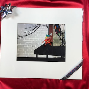 Puede incluir: Un cel de animación enmarcado que presenta un personaje de dibujos animados con un traje rojo y naranja de pie en una plataforma. El personaje lleva un casco azul y blanco y sostiene un bastón. El fondo es una pared de ladrillo gris con cables negros y un foco.