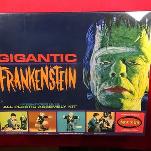 Peut inclure: Boîte de kit d'assemblage en plastique Gigantic Frankenstein. La boîte présente une grande image du visage de Frankenstein en vert, bleu et jaune. Le titre "GIGANTIC FRANKENSTEIN" est en orange et violet. Comprend le logo Moebius Models.