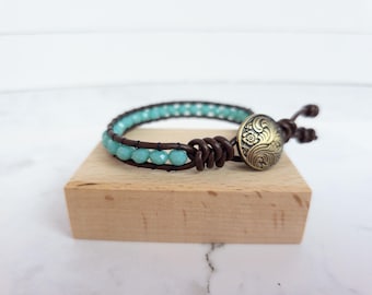 May include: A wrap bracelet featuring turquoise beads and dark brown leather. The bracelet has a decorative bronze-colored button clasp. The bracelet is displayed on a small wooden block.