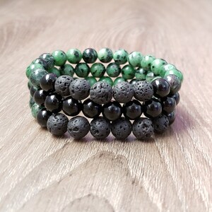 May include: Three layered bracelets with green, black, and dark grey beads. The green beads are a mottled green and black color. The black beads are smooth and shiny. The dark grey beads are rough and porous.