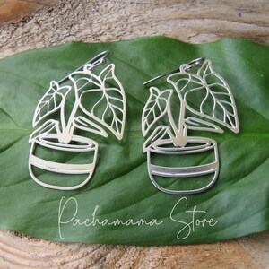 May include: A pair of silver plant earrings. Each earring shows a plant design with leaves and a pot. The earrings are displayed on a large green leaf, with the text "Pachamama Store" in cursive.