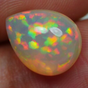 Pode incluir: Uma gema de opala em forma de lágrima com uma base branca e um jogo de cores colorido, incluindo verde, laranja e vermelho.