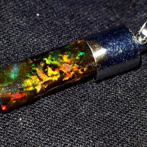 Pode incluir: Um pingente de prata com uma pedra de opala retangular. A opala tem uma base escura com flashes de verde, amarelo e vermelho.