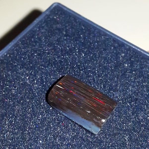 May include: A rectangular black opal gemstone, polished to a high sheen, displaying flashes of red and blue. The opal is set against a dark blue, textured surface, highlighting its iridescent colours. Presented in a dark blue box.