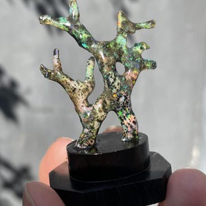 Puede incluir: Una escultura artesanal única que se asemeja a una rama de árbol, mostrando un espectro de colores que incluye verde, dorado y toques de rosa. La escultura está montada sobre una base hexagonal negra, creando un contraste llamativo. Una pieza decorativa cautivadora.