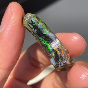 Op de afbeelding: Een levendige, ruwe opaal edelsteen toont een spectrum aan kleuren, waaronder groen, zwart en oranje. De opaal wordt tussen vingers gehouden, wat de natuurlijke, ruwe textuur en het iriserende kleurenspel benadrukt. De edelsteen is een uniek stuk natuurlijke sieraden.