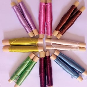 May include: Eight spools of thread wound around wooden sticks. The spools are arranged in a starburst pattern. The thread colours are purple, pink, brown, beige, yellow, green, burgundy, and blue.