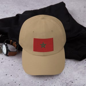 Puede incluir: Una gorra de béisbol de color beige con una bandera roja de Marruecos bordada con una estrella verde de cinco puntas.