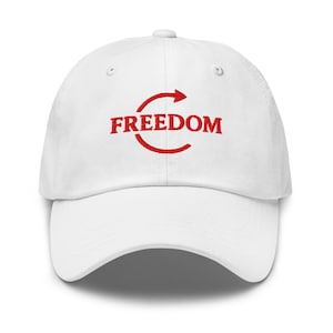 May include: White baseball cap with the word "FREEDOM" in red, arched above a red arrow. The cap has a curved brim and a fabric strap closure on the back.