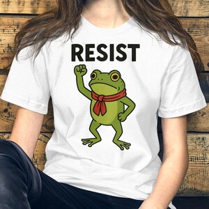 May include: White t-shirt with the word "RESIST" in bold black letters. A green cartoon frog with a red scarf is raising its fist. The frog is centered on the shirt.