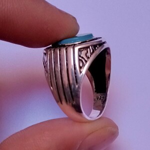 Peut inclure: Une bague en argent avec une pierre turquoise sertie au centre. La bague a un design complexe avec une bande texturée.