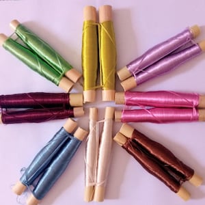 May include: Eight spools of thread in various colours, including green, yellow, purple, pink, maroon, blue, white, and brown, are arranged in a starburst pattern on a white surface.