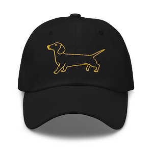 Könnte beinhalten: Schwarze Baseballkappe mit gelber Umrisszeichnung eines Dackelhundes. Die Kappe hat einen gebogenen Schirm und einen verstellbaren Riemen. Die Hundeillustration ist mittig auf der Vorderseite platziert.