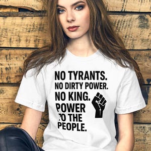 Peut inclure: T-shirt blanc avec le texte "NO TYRANTS. NO DIRTY POWER. NO KING. POWER TO THE PEOPLE." et un poing noir. Le t-shirt est porté par une personne aux longs cheveux bruns.