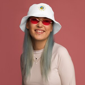 Puede incluir: Una mujer con el pelo azul lleva una gorra de pescador blanca con un diseño de sol amarillo y pelota de playa azul. También lleva gafas de sol rojas en forma de corazón.