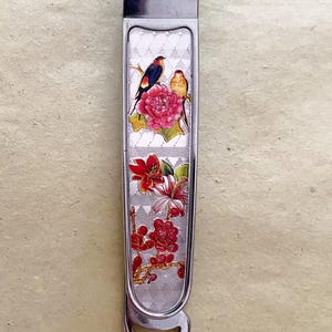 Può includere: Tagliaunghie argentato con un design decorativo floreale e di uccelli. Le forbici per unghie presentano un'illustrazione dettagliata di fiori rossi e due uccelli appollaiati su un ramo, su uno sfondo strutturato.