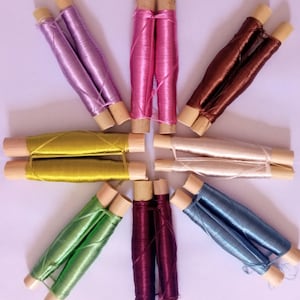 May include: Eight spools of thread wound on wooden bobbins. The thread colours are green, burgundy, light blue, cream, gold, brown, pink, and lavender.