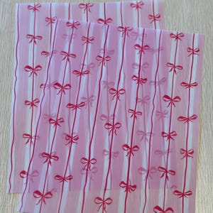 May include: Two sheets of pink wrapping paper with a repeating pattern of red bows and vertical stripes. The bows are evenly spaced along the red stripes, creating a delicate and feminine design.