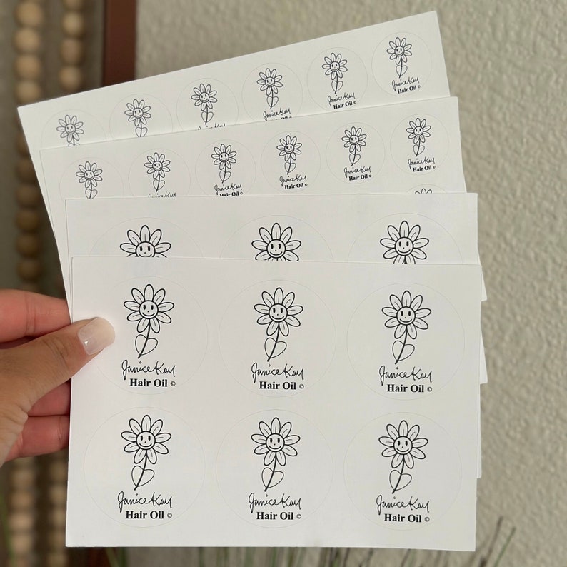 May include: A sheet of white stickers with a black and white illustration of a flower with a smiley face. The text "Janice Kay Hair Oil" is written below the flower.