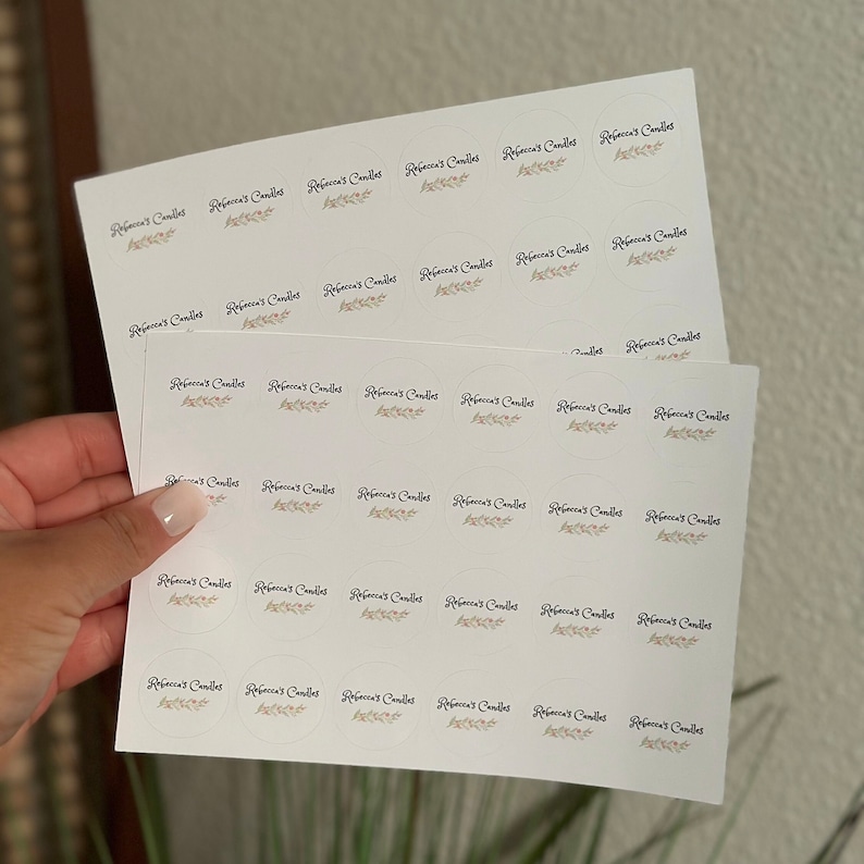 May include: A sheet of white sticker labels with the text "Rebecca's Candles" printed in black cursive font with a small brown leaf design. The labels are arranged in rows and columns.