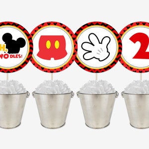 May include: Four red, yellow, and black circular Mickey Mouse themed party decorations with a white background. The decorations feature Mickey Mouse's head, shorts, hand, and the number 2. The decorations are on sticks and placed in silver buckets with white tissue paper.