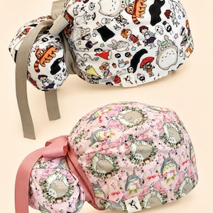 Puede incluir: Dos gorros de quirófano con bolsa para coleta y cintas. El gorro superior es blanco con un estampado de personajes de dibujos animados multicolores. El gorro inferior es rosa con un patrón repetitivo de criaturas grises, coronas florales y pequeños lazos rosas.