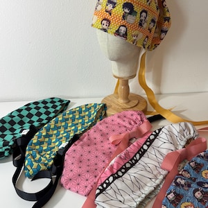Op de afbeelding: Een collectie van zes stoffen operatiemutsen met diverse anime-geïnspireerde patronen, waaronder een gele muts met karakterprints op een etalagepophoofd, en vijf andere in groene, blauwe, roze en witte geometrische ontwerpen met bevestigde linten.