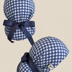 Puede incluir: Dos gorros quirúrgicos a cuadros azules y blancos con lazos azul marino. Los gorros son redondos y fruncidos en la parte trasera, con lazos largos. Un gorro tiene una pequeña etiqueta blanca con un logotipo negro.