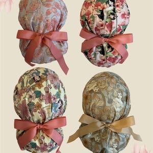 May include: Four scrub caps featuring floral patterns and cartoon character illustrations. Each cap has a gathered back secured with a tied ribbon. The designs include various pastel and vibrant color palettes with pink, yellow, and muted blue tones.