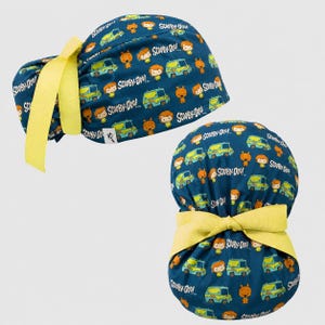 Puede incluir: Un gorro quirúrgico azul oscuro con un patrón repetitivo de personajes de Scooby-Doo y la furgoneta Mystery Machine, acentuado con una cinta amarilla brillante. La tela muestra el logotipo de Scooby-Doo en texto blanco en todo el diseño.