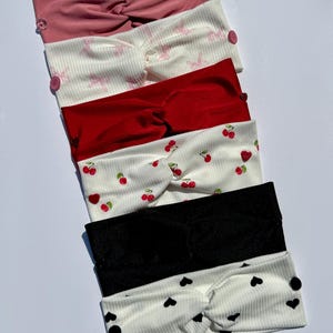 Puede incluir: Una colección de seis diademas en varios colores y diseños. Las diademas incluyen colores lisos como el rosa, el rojo y el negro, así como diseños estampados con cerezas, corazones y conejos. Cada diadema tiene un diseño anudado y botones decorativos.