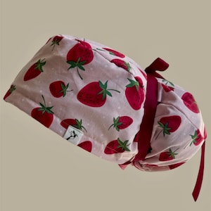 Puede incluir: Un gorro quirúrgico rosa claro con un estampado de fresas rojas grandes con hojas verdes. El gorro tiene una cinta burdeos y una pequeña etiqueta blanca con un logotipo. El fondo es de color beige claro.