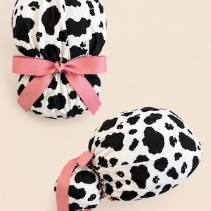 May include: A fabric hair accessory featuring a black and white cow print pattern. The accessory has a rounded, gathered shape and is accented with a solid dusty rose pink ribbon tied into a bow at the center, with two long ribbon tails hanging down.