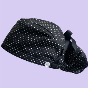 Puede incluir: Un gorro quirúrgico negro con estampado de lunares blancos, un botón blanco en el lateral para sujetar accesorios y una cinta negra en la parte posterior para ajustar el ajuste. La tela está fruncida en la parte trasera para diversas longitudes de pelo.
