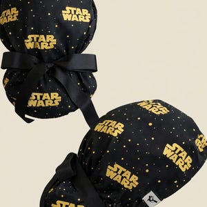 May include: Black scrub cap with the Star Wars logo in gold. The cap has a black ribbon and is patterned with gold dots. The Star Wars logo is repeated across the fabric.