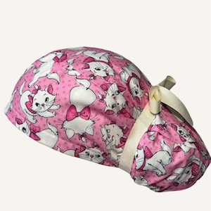 Puede incluir: Un gorro quirúrgico rosa con un estampado de gatos blancos de dibujos animados con lazos rosas. El gorro tiene una banda blanca y un lazo color crema en el lateral. El fondo tiene un estampado de lunares.
