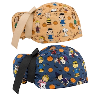 May include: Two scrub caps featuring Peanuts characters. The top cap is tan with pumpkins and autumn leaves. The bottom cap is blue with pumpkins, bats, and characters in Halloween costumes. Both caps have a solid color ribbon tie at the back.