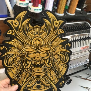 Puede incluir: Un parche bordado en oro que representa un casco de samurai con una expresión feroz. El parche está sobre un fondo negro y está siendo sostenido por una mano.