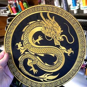 Peut inclure: Un écusson rond noir avec un motif de dragon brodé en or. Le dragon est représenté dans une pose incurvée, avec des détails complexes. Une bordure dorée de clé grecque encadre le dragon. L'écusson est tenu par une main, avec des bobines de fil en arrière-plan.