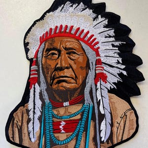 Peut inclure: Patch brodé représentant un homme amérindien portant une coiffe traditionnelle avec des plumes. L'homme a une expression sérieuse et porte un collier de perles. Le patch est sur un fond noir.