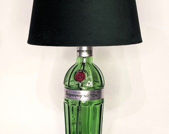 Può includere: Una lampada a forma di bottiglia di vetro verde con un paralume in tessuto nero. La bottiglia è etichettata "Tanqueray No. Ten" e ha un sigillo rosso. La lampada ha un cavo e un interruttore neri.