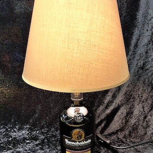 Op de afbeelding: Een tafellamp met een voet van een donkerglazen fles Bunnahabhain 12 jaar oude Islay single malt Scotch whisky, bekroond met een beige stoffen trommelkap. Het etiket op de fles bevat de tekst Bunnahabhain, 12 en 1881.